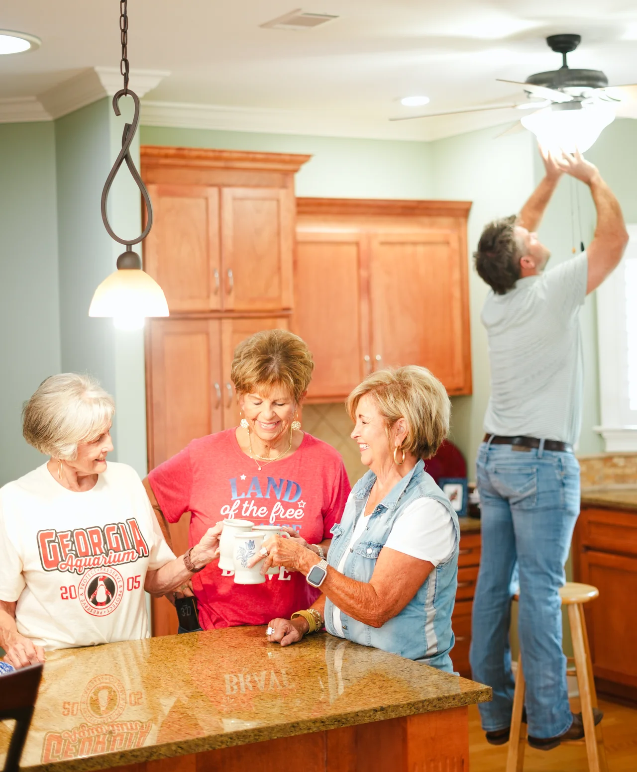 A man changing a ceiling light as three women chat at a kitchen counter.